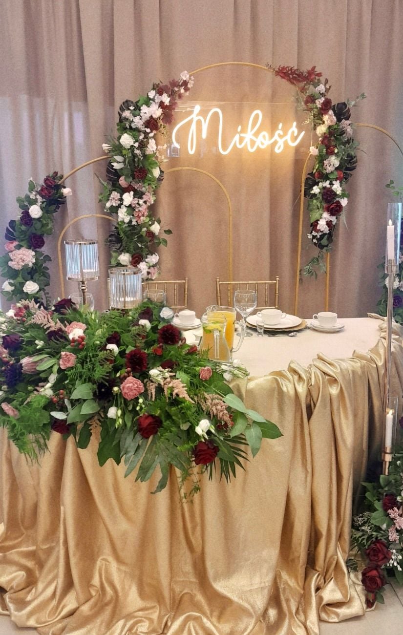 Nakryty stół weselny dla Państwa Młodych ze złotym obrusem i kwiatową kompozycja i neonem Miłość